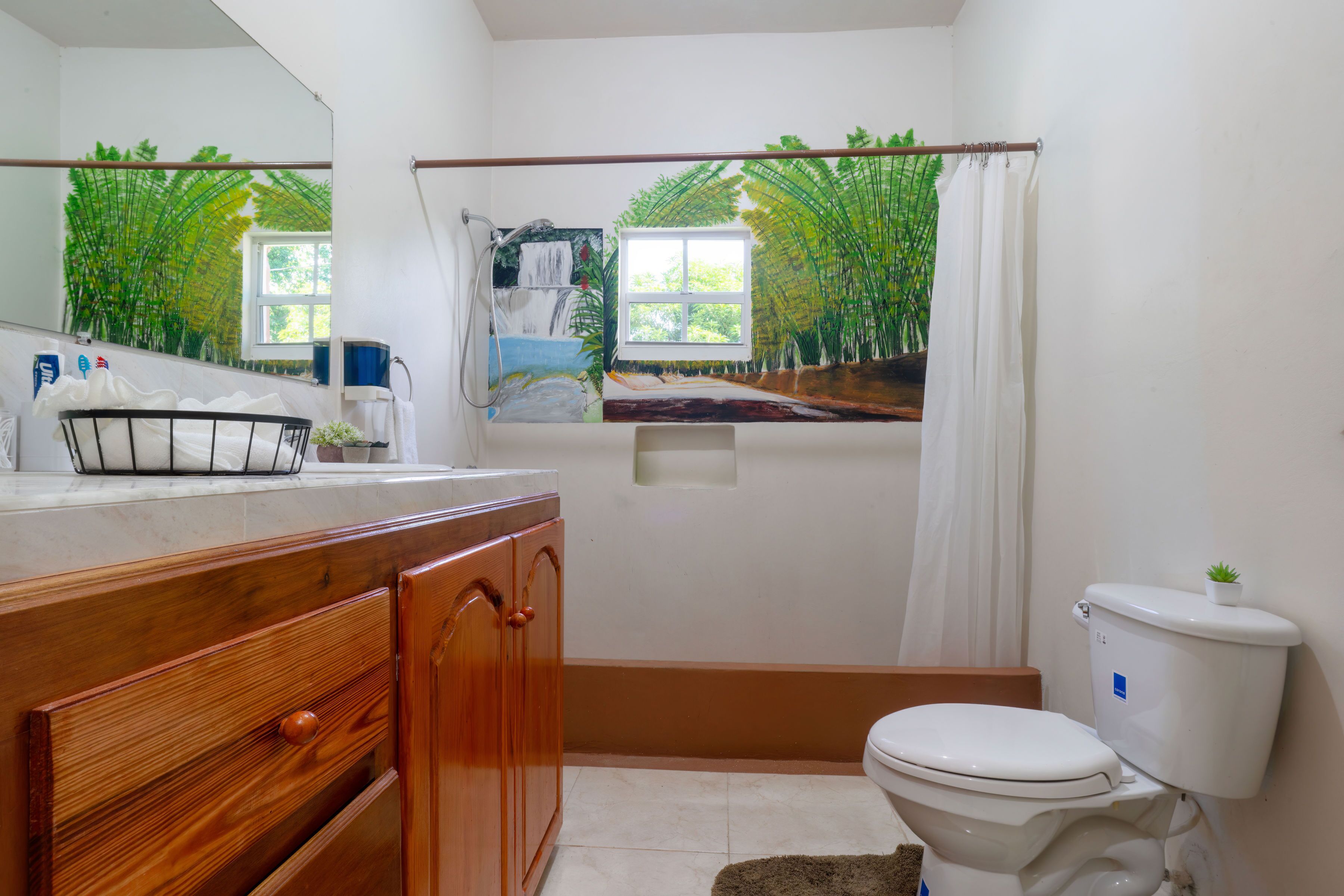 A bathroom with a wooden vanity, a toilet, a shower curtain, and a window with a view of bamboo plants.