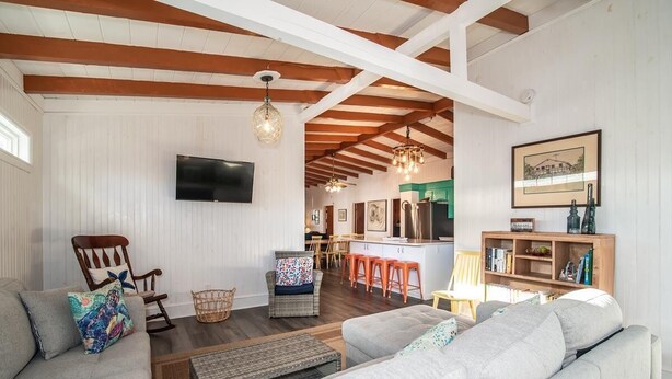 Ein geräumiges Wohnzimmer mit einer großen Couch, einem Korbstuhl und einem Essbereich mit orangefarbenen Hocker. Ein Fernseher ist an der Wand befestigt, und es gibt ein Bücherregal mit diversen Gegenständen.