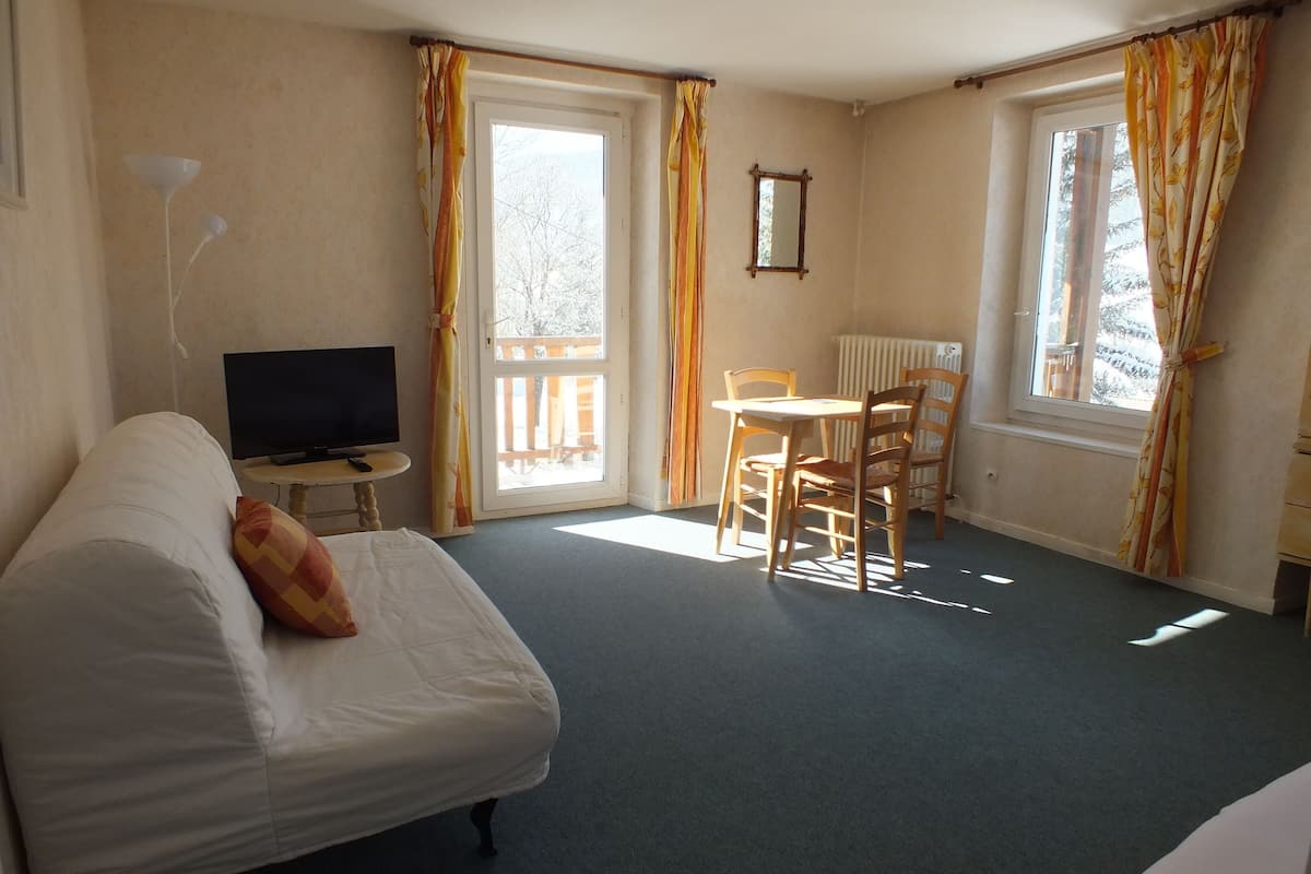 A hotel room with a bed, a TV, a small table with chairs, and large windows with curtains.
