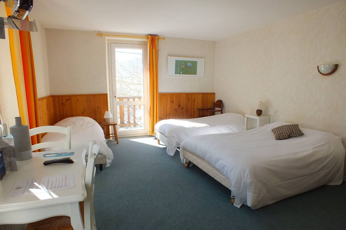 A hotel room with two single beds, a small table with a chair, a desk with a chair, a window with curtains, and a framed picture on the wall.