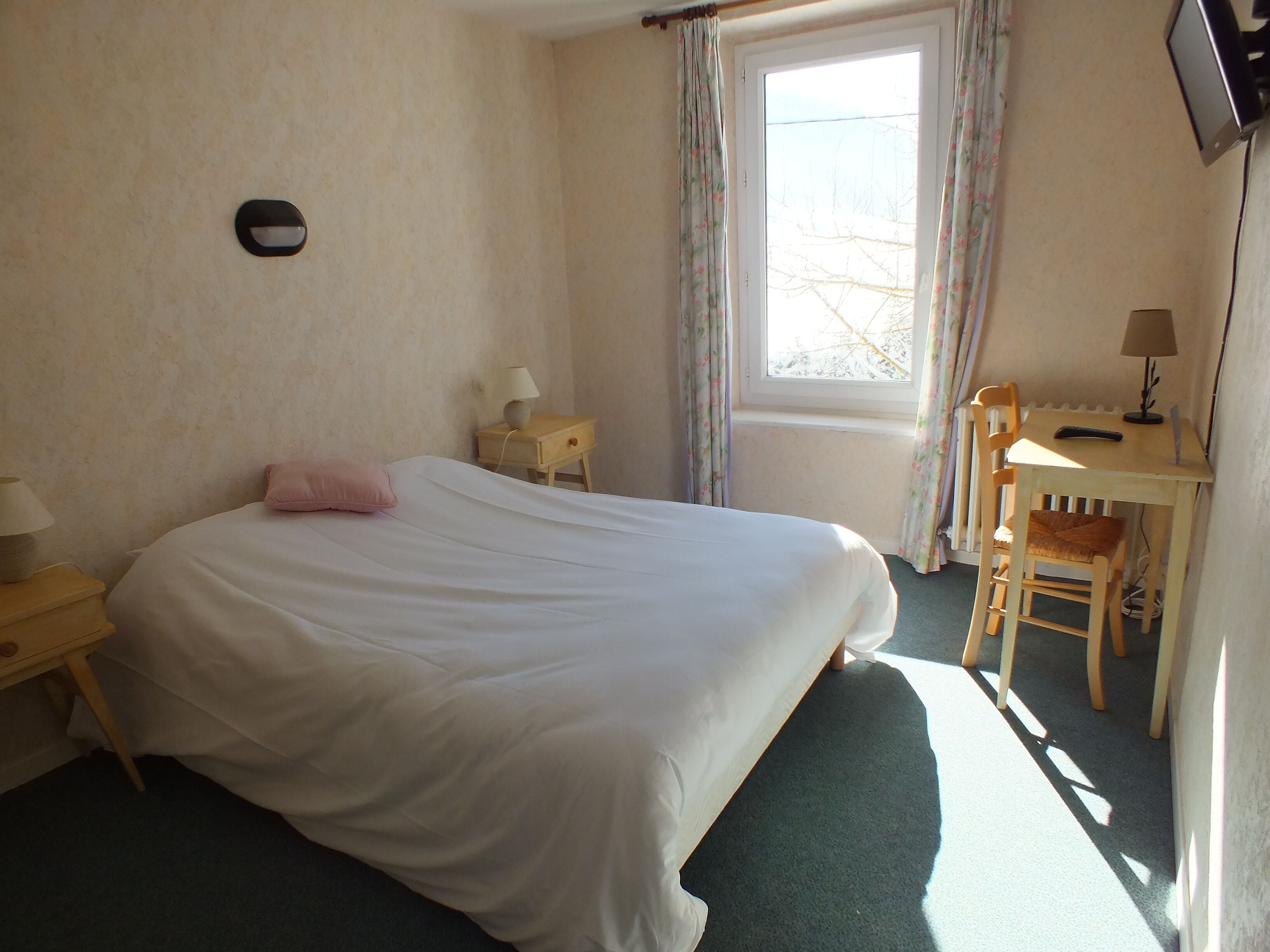 A bedroom with a bed, a desk, a chair, a nightstand, a lamp, and a window with curtains.