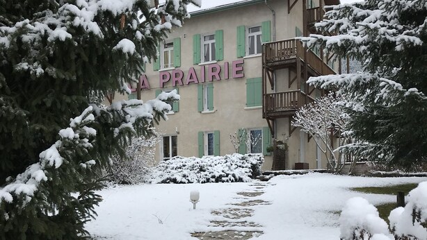 Un edificio con un letrero que dice "LA PRAIRIE", rodeado de árboles cubiertos de nieve y un camino de piedra.