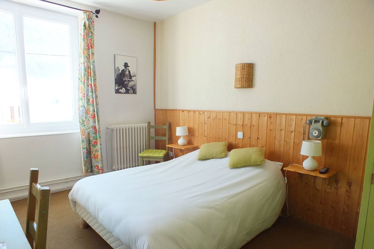 A hotel room with a large bed, two bedside lamps, a wooden headboard, a radiator, a chair, a table, and a telephone.