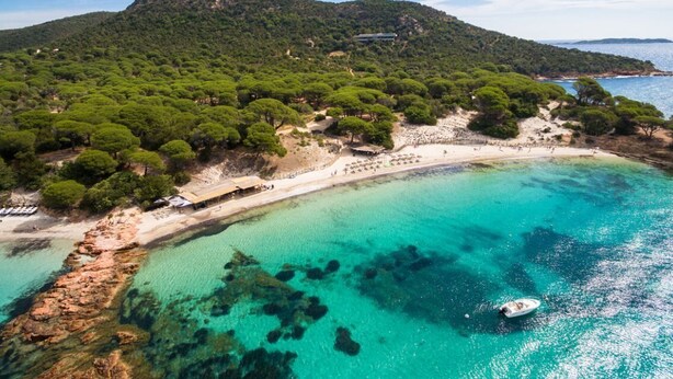 Vue aérienne d’une zone côtière avec une plage, des eaux turquoise et limpides, des affleurements rocheux et des collines verdoyantes et densément boisées.