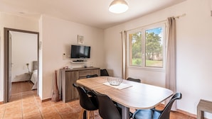 Un coin repas avec une table en bois, des chaises noires, un téléviseur posé sur un meuble et une fenêtre avec des rideaux.