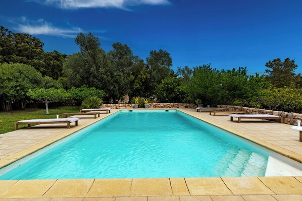 A rectangular pool with a stone border, surrounded by lounge chairs and a stone wall.