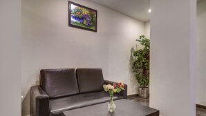 Um hall de hotel moderno com um sofá de couro, um vaso de flores sobre uma mesa, uma planta em vaso grande e um quadro na parede.