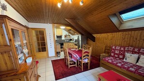 A cozy room with wooden walls and a sloped ceiling, featuring a dining area with a table and chairs, a sofa with patterned cushions, and a small bookshelf. - Châtel - Roitet : 3P apartment, quiet, breathtaking view in Chalet Lou P'nots  (Châtel)