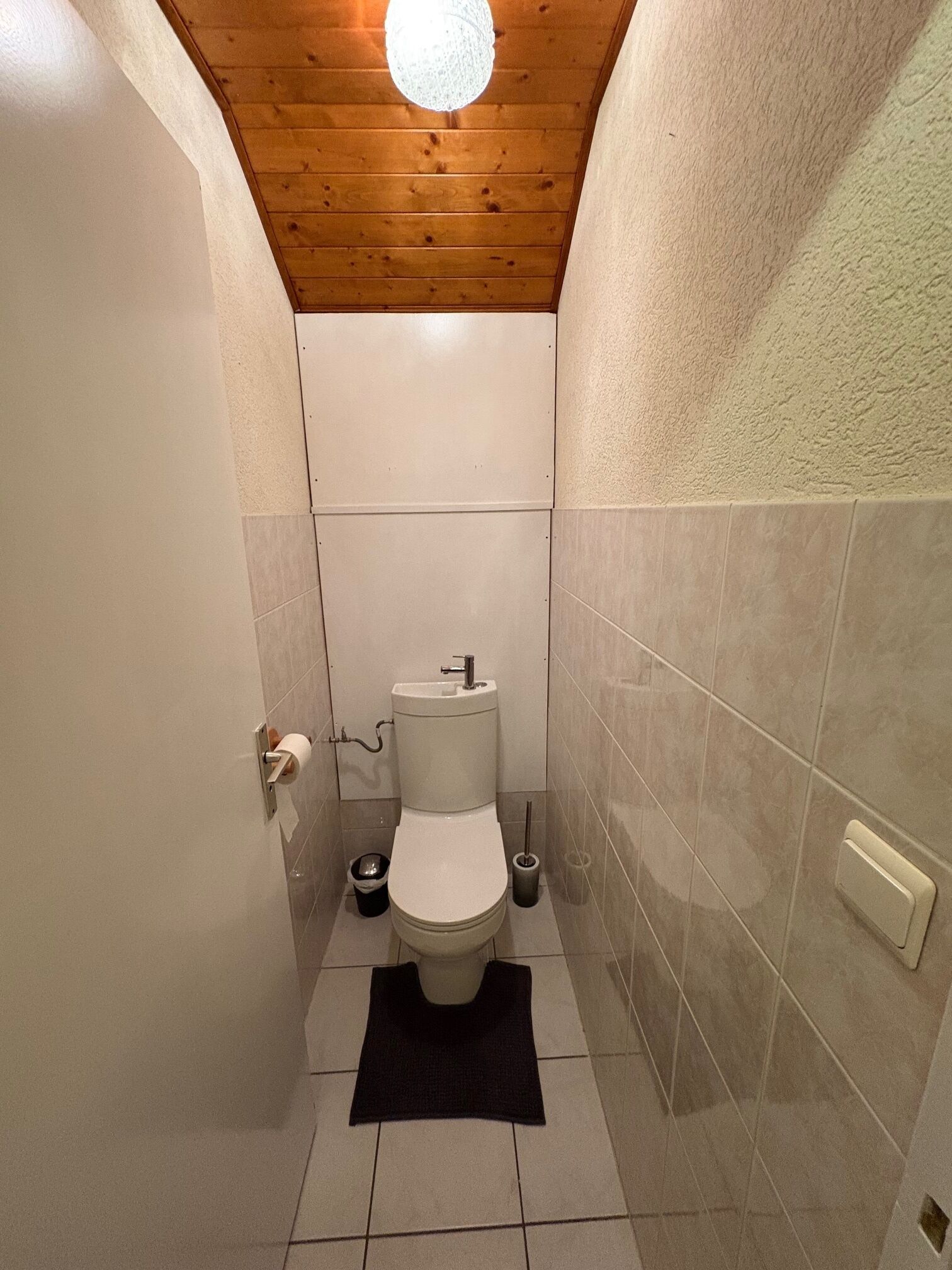 A small, narrow bathroom with a wooden ceiling, white tiles, a toilet, and a black rug.