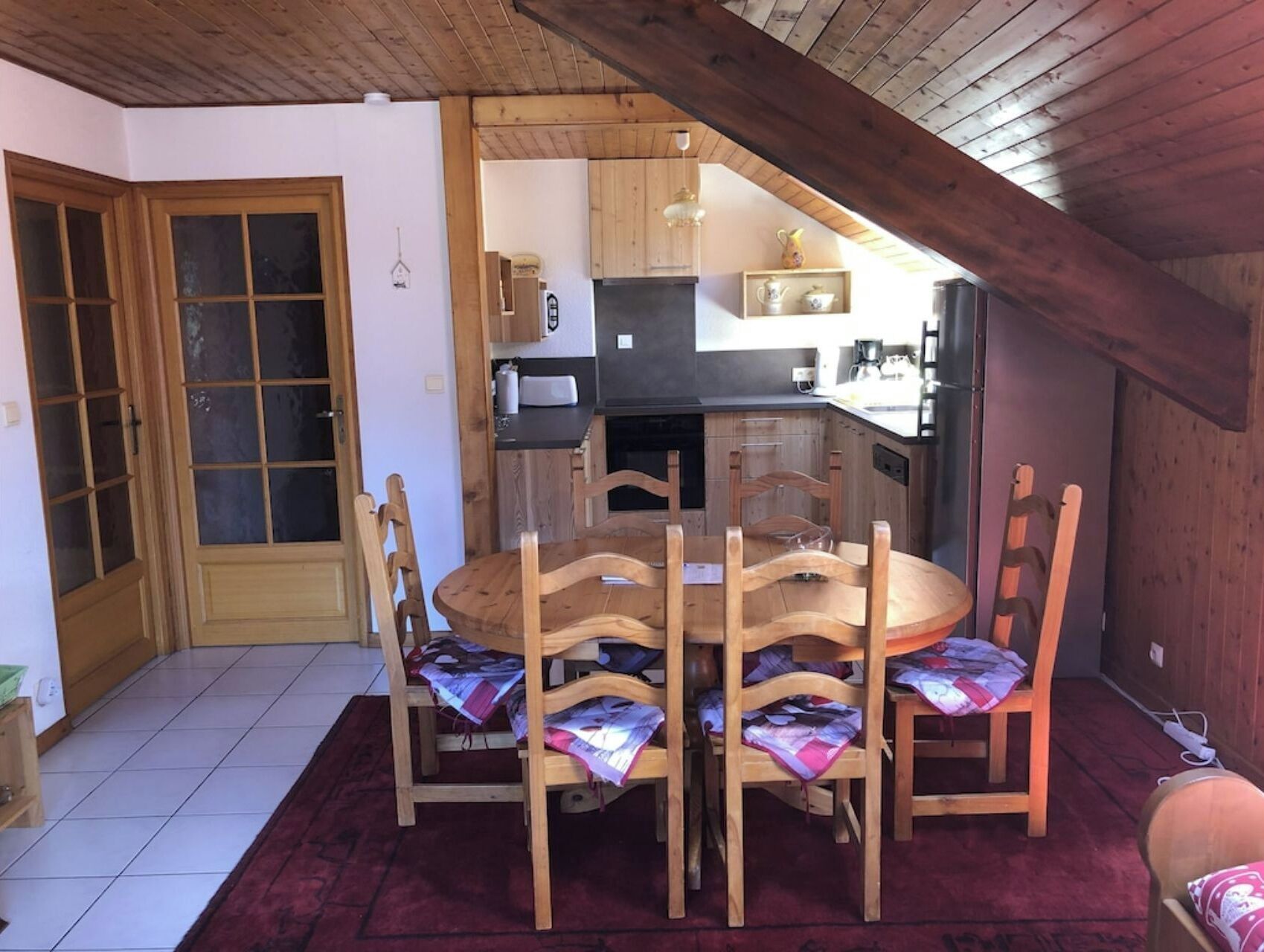 A dining area with a round wooden table and six chairs, a red carpet, and a kitchen in the background.