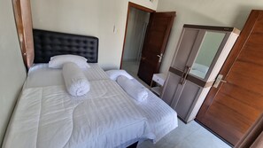 Una cama bien hecha con sábanas blancas y dos toallas enrolladas, un cabecero oscuro, un armario de madera y una mesita de noche pequeña.