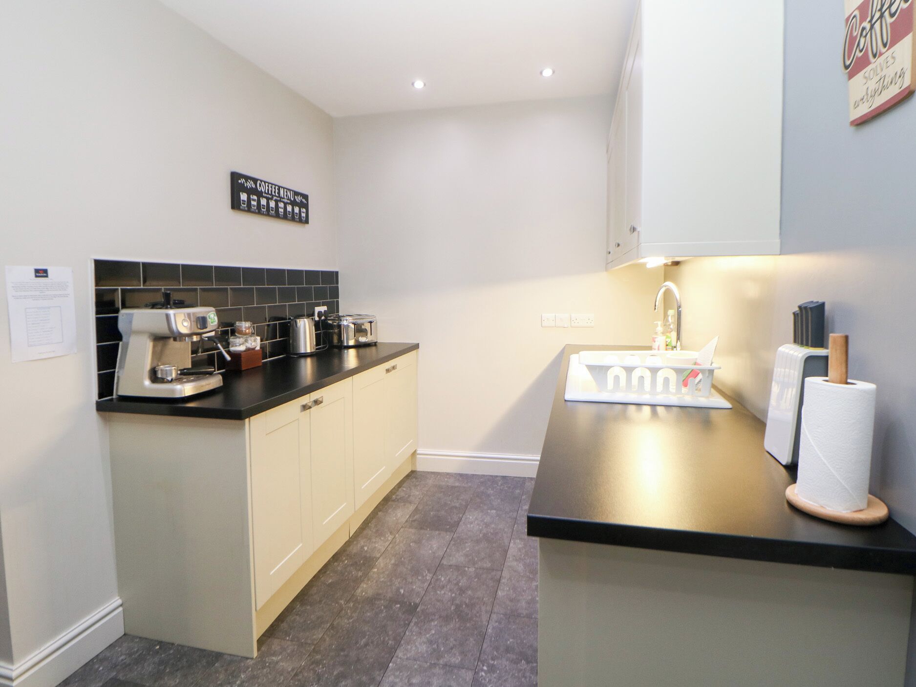 A modern kitchen with a coffee machine, toaster, and kettle on the counter, a sink with a paper towel holder, and a sign indicating a coffee menu.