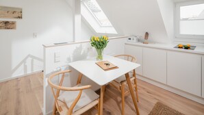 A small, modern kitchen with a white table, two wooden chairs, a vase with yellow flowers, and a small radio on the counter. - House Salz&Sand // Apartment Lüttje Tide (Norderney)