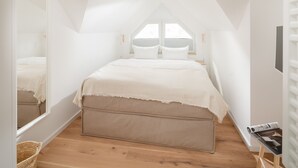 A bedroom with a high ceiling, a bed with a beige cover, a wooden floor, and a large mirror. - House Salz&Sand // Apartment Lüttje Tide (Norderney)