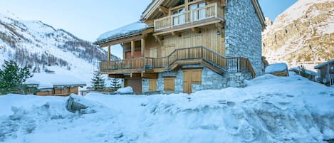 A multi-story wooden chalet with stone walls, a balcony, and a snow-covered landscape.