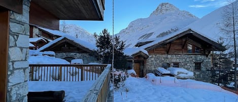 A snow-covered mountain lodge with stone walls and wooden beams, featuring a wooden deck and a view of the surrounding snowy landscape.