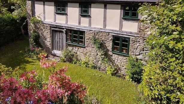 A two-story house with a stone facade, multiple windows, and a garden with flowering plants. - Luxurious Cottage with Lovely Views in Rural Location (Norton Presteigne)