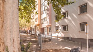 A courtyard with a tree, a metal fence, and a building with multiple windows and a trash bin. - Central, modern designer appartment in downtown Stuttgart (Stuttgart)