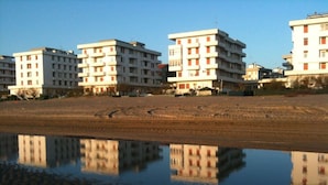 Una hilera de edificios de varios pisos con balcones, una playa arenosa al frente y una masa de agua tranquila que refleja los edificios.