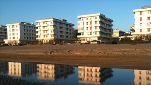 Una hilera de edificios de varios pisos con balcones, una playa arenosa al frente y una masa de agua tranquila que refleja los edificios.