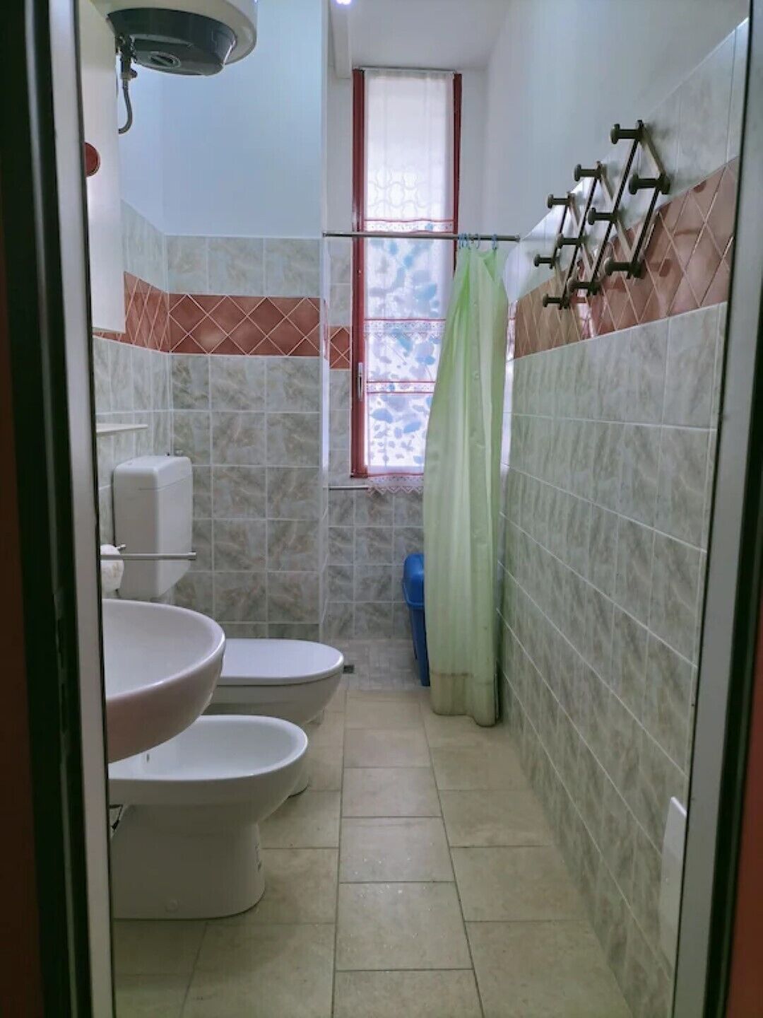Une petite salle de bain avec deux toilettes, un lavabo, une fenêtre avec un rideau et des murs carrelés.