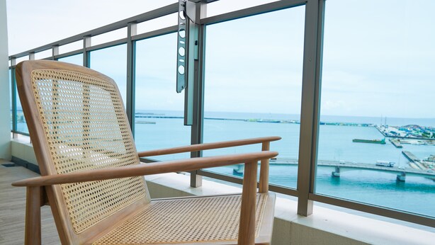 A wooden chair with a woven backrest and armrests, positioned by a large window overlooking a body of water and a bridge.