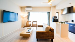 A modern living room with a sofa, a coffee table, and a TV. There is a kitchen area with a microwave and a kettle.