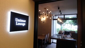 A modern interior with a sign reading "Umimae Lounge," a dining area with a wooden table and chairs, and a hanging light fixture.