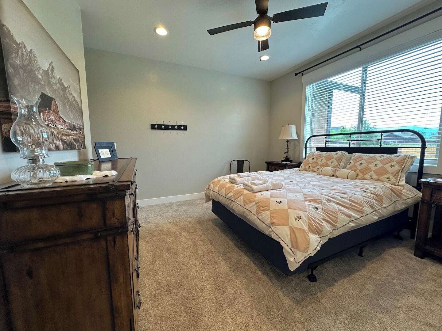 A bedroom with a bed, a dresser, a ceiling fan, a window with blinds, and a wall-mounted light switch.