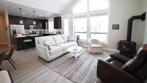 A modern living room with a sofa, armchair, and coffee table, a kitchen with dark cabinets and a stove, and large windows providing natural light. - Luxurious Family Home in Driggs - Hot Tub (Driggs)