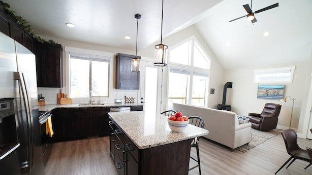 A modern kitchen with dark cabinets, a granite countertop, and a central island with a bowl of fruit. There is a dining area with a sofa and chairs, and a fireplace. - Luxurious Family Home in Driggs - Hot Tub (Driggs)