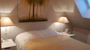 Un dormitorio con cama, dos lámparas de noche, una mesita de noche, un cuadro de árboles y un tragaluz.