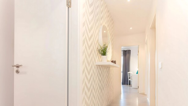 A hallway with a white door, a chevron-patterned wall, a plant on a floating shelf, and a glimpse of a room with a chair and a desk.