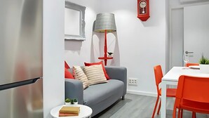 A modern living room with a grey sofa, red cushions, a small round table with a book, a red lamp, and a dining area with a white table and orange chairs.