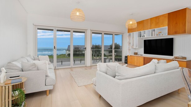 A modern living room with a large TV, two sofas, and a view of the ocean through glass doors. - Prime Position -  Family Getaway (Gerroa)