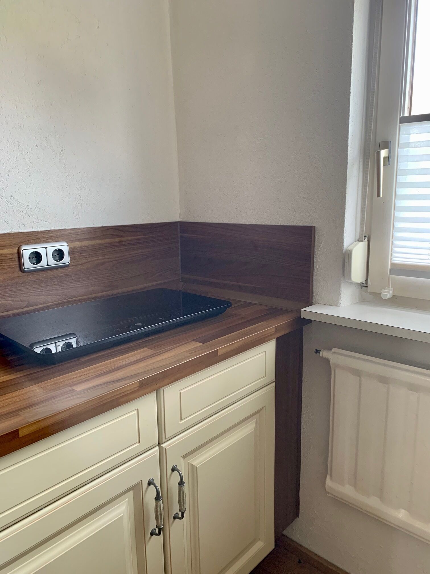 Une cuisine dotée d’un plan de travail en bois, de deux prises électriques, d’une fenêtre avec stores et d’un radiateur blanc.