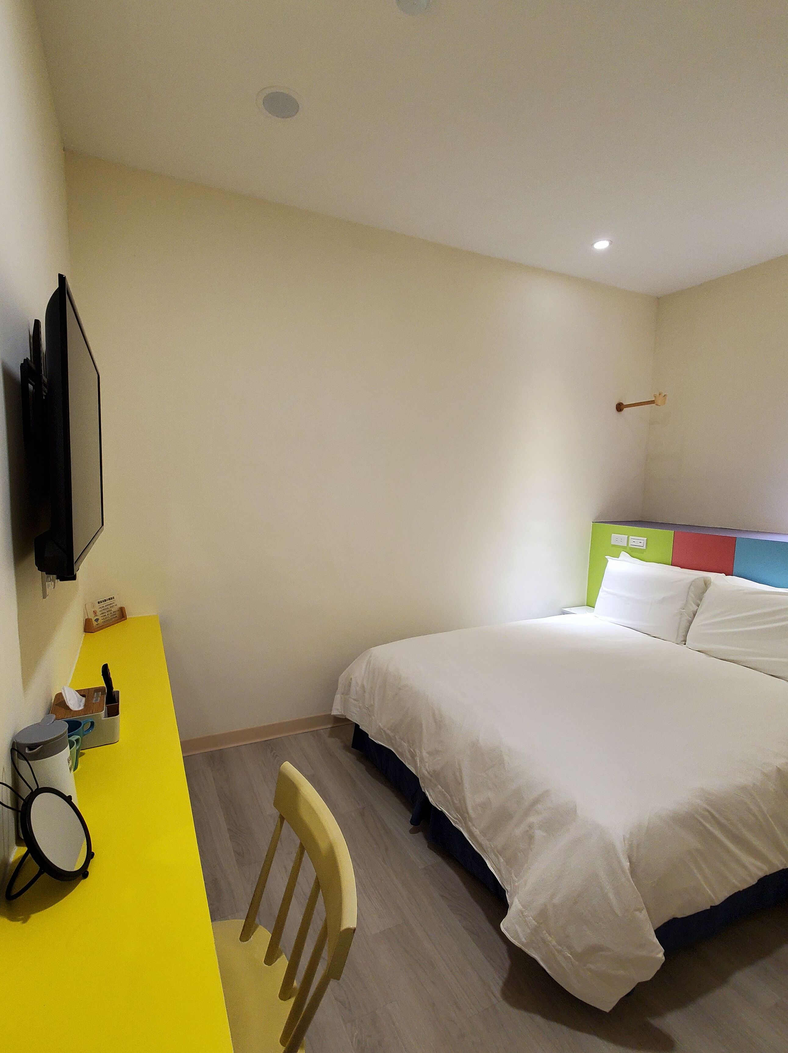 A hotel room with a bed, a desk with a chair, a television, and a wall-mounted shelf.