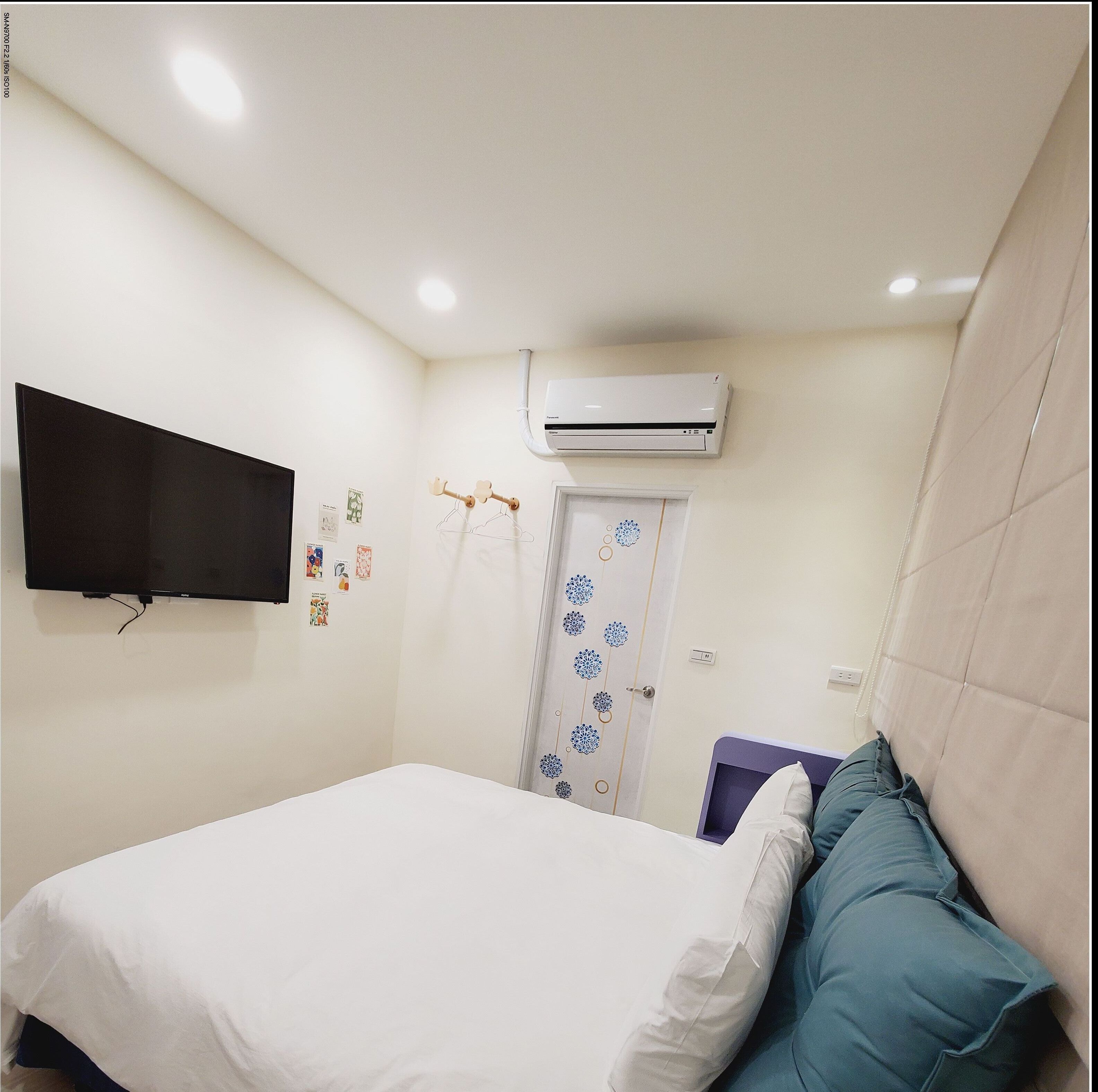 A bedroom with a bed, a TV mounted on the wall, an air conditioning unit, and a door with a decorative pattern.
