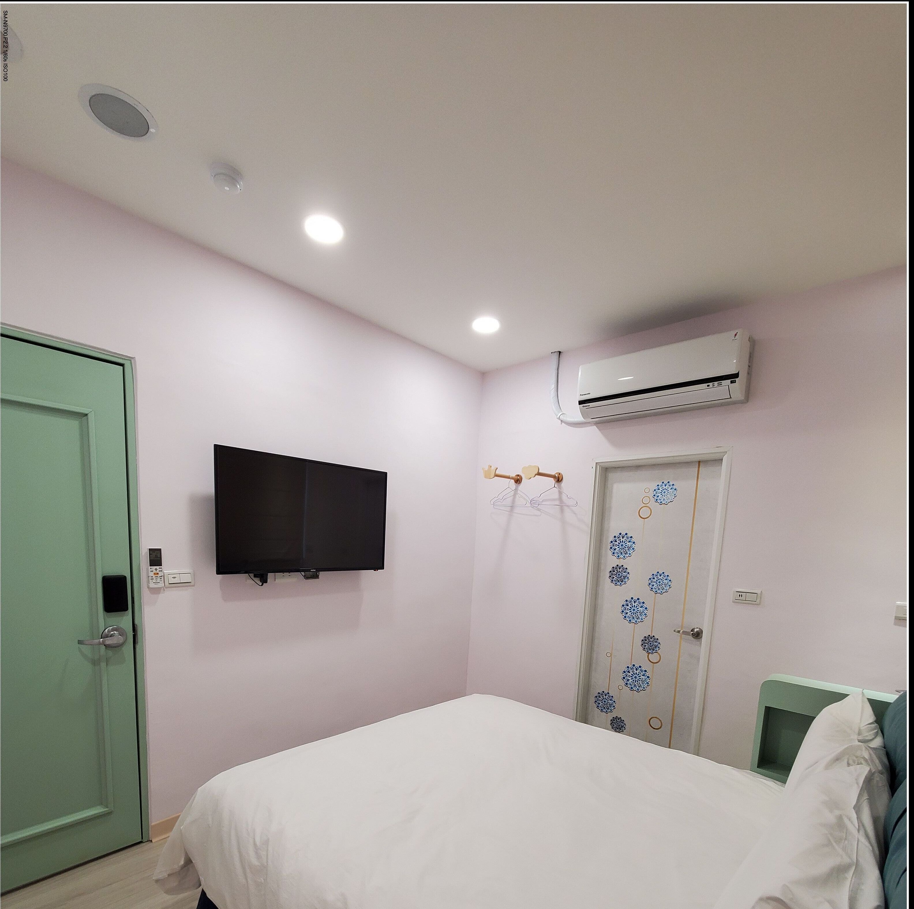 Un dormitorio con cama, televisor montado en la pared, unidad de aire acondicionado, una puerta con diseño decorativo y una puerta verde.