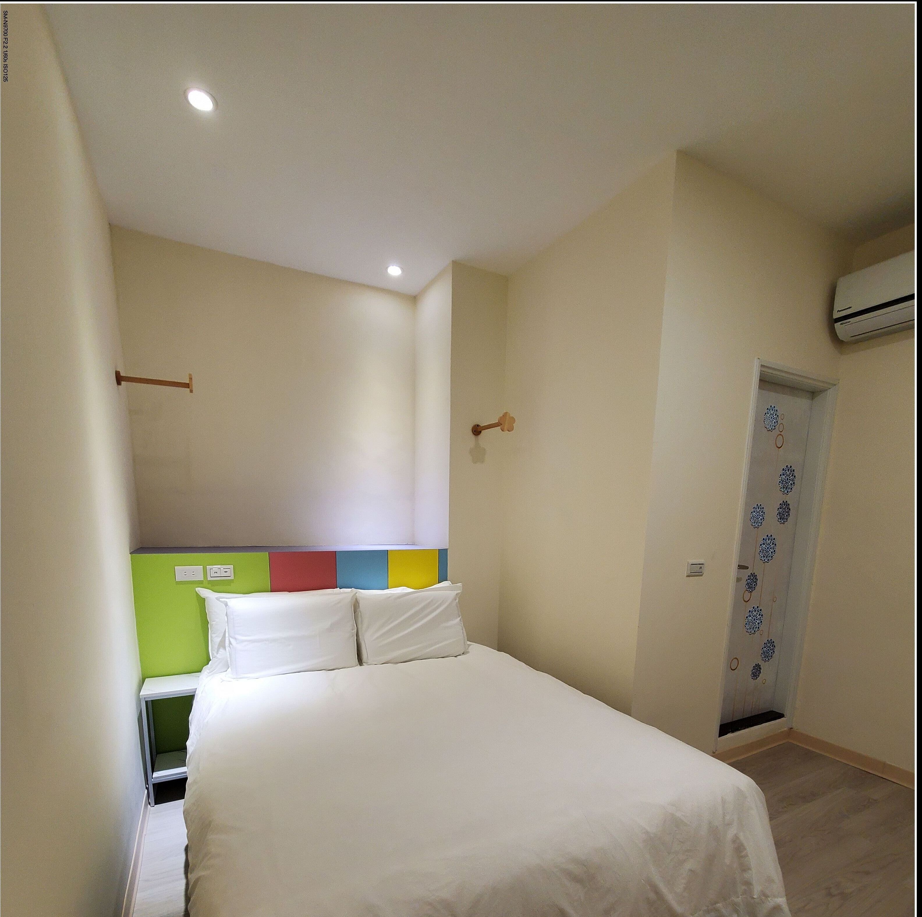 A hotel room with a bed, a bedside table, a wall-mounted air conditioner, and a door with a decorative pattern.