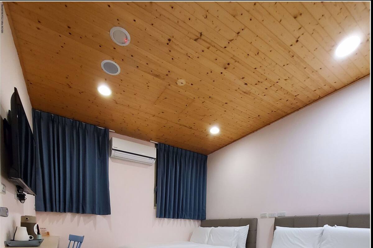A bedroom with a wooden ceiling, a bed, a desk with a chair, and an air conditioning unit.