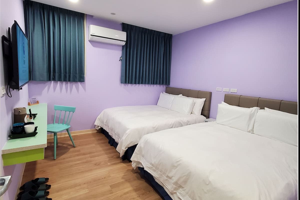 A hotel room with two beds, a desk with a chair, an air conditioner, and a TV mounted on the wall.