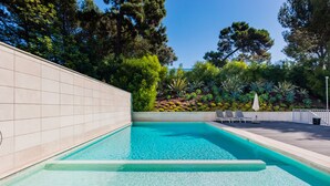 A rectangular swimming pool with clear blue water, surrounded by a tiled wall and a landscaped area with various plants and trees. - Santo Amaro Beach Apartment (Oeiras)