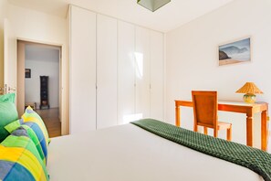 A bedroom with a bed, a desk, and a chair. There is a wardrobe, a picture on the wall, and a lamp on the desk.