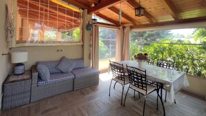 Una habitación acogedora con techo de madera, un sofá con cojines estampados, una mesa de comedor con sillas y grandes ventanales con vistas a la vegetación.