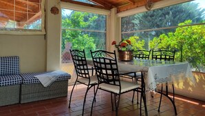Un solárium con techo de madera, una mesa con sillas, un banco acolchado y vistas al exterior con vegetación.