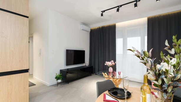A modern living room with a flat-screen TV, a dining table with chairs, a sofa, and a large window with curtains.