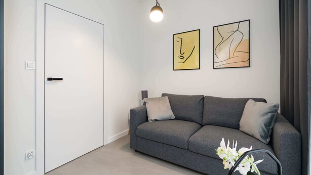 A modern living room with a grey sofa, two abstract paintings, a white door, and a small table with a lamp.