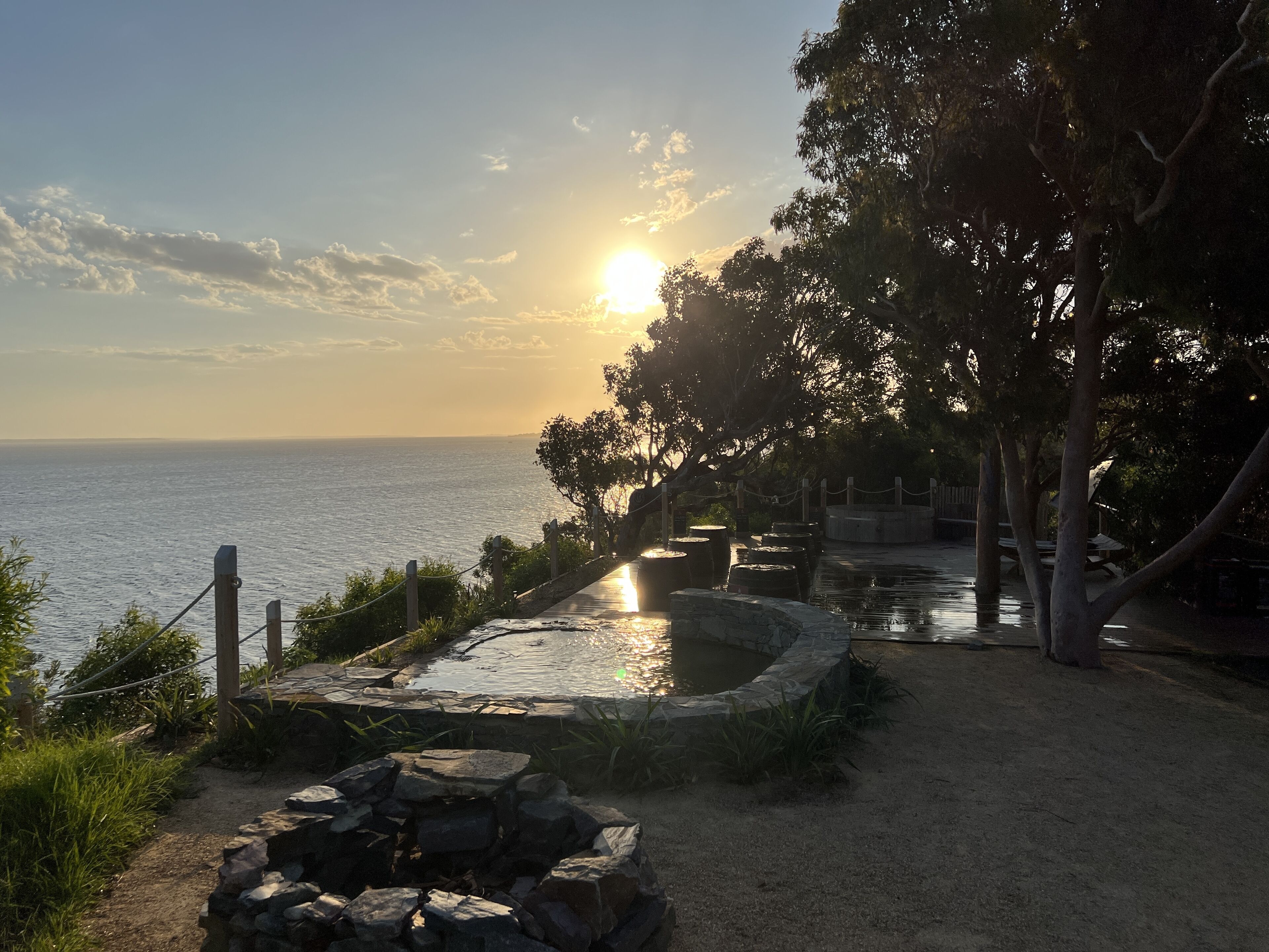 A stone pathway leading to a small pond with a wooden fence and trees on one side, and a view of the sea on the other.