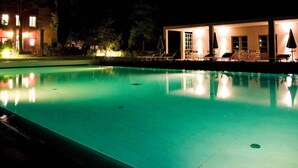 En pool för nattbad med klart vatten, omgiven av solstolar och parasoller, och en byggnad med upplysta fönster i bakgrunden.